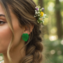 Close-up van een vrouw met gevlochten haar en bloemen in het haar, die een groene oorsteker in de vorm van een Monstera (gatenplant) blad draagt in een bosrijke omgeving.
