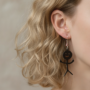 Close-up van het oor van een vrouw met golvend blond haar die een zwarte, druppelvormige oorbel draagt in de vorm van een vrolijke stickman met een glimlachend gezicht.