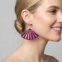 Close-up van een vrolijk, blond model met een grote paarse oorbel in waaiervorm met uitgesneden spijlen. De oorbel is 60 mm breed en hangt aan een zilveren oorhaakje.