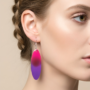 Close-up van een vrouw die een lange, ovale handgemaakte resin oorbel draagt met een kleurverloop van fuchsia roze naar paars.