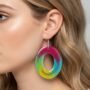 Zijaanzicht van een blond model dat een grote ovale epoxy statement oorbel draagt met een abstract en kleurrijk patroon van roze, groen en blauw op een neutrale achtergrond.