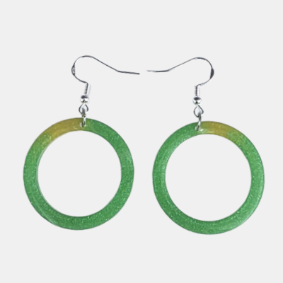 Paar handgemaakte ronde oorbellen van groene epoxy met fijne glitters en een subtiel geel kleuraccent aan de bovenzijde op een lichte achtergrond.