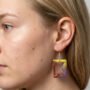 Close-up van een model met blond haar die handgemaakte rechthoekige epoxy oorbellen in geel en paars draagt