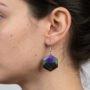 Close-up foto van een donkerharig vrouwelijk model in profiel, die een handgemaakte zeshoekige resin oorbel draagt met een uniek swirlpatroon in diep paars, zwart en emerald groen glitter.
