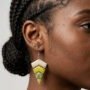 Close-up van een model met donkere huid en vlechtjes, die gelaagde geometrische epoxy oorbellen draagt met chevronvormen in wit, geel en groen